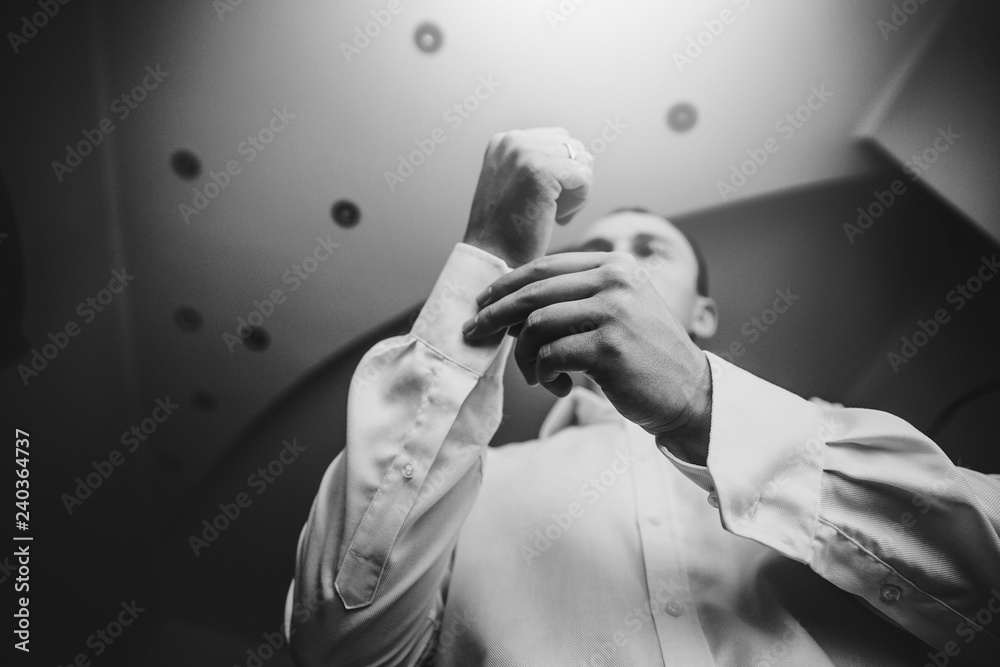 Stylish groom putting on white shirt, getting ready in the morning near window in soft light, before wedding ceremony. Bottom top view