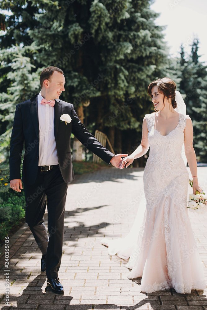 Gorgeous bride in amazing gown and stylish groom walking and smiling in sunny city street. Beautiful happy wedding couple enjoying time in sunny outdoors. Romantic moments