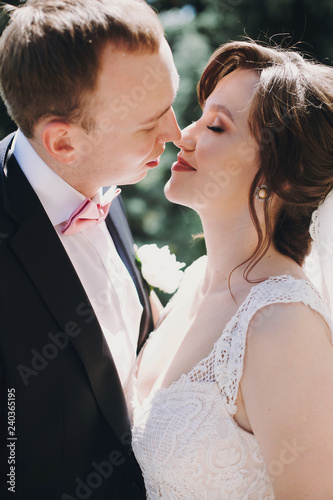 Gorgeous bride in amazing gown and stylish groom gently kissing in sunny park. Beautiful happy wedding couple enjoying time and embracing outdoors. Romantic sensual moment