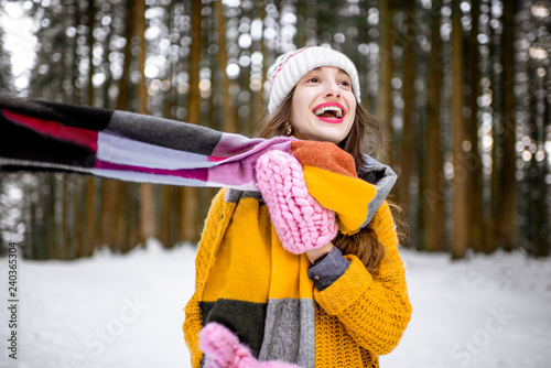 Portrait of a young playful woman dressed in bright winter clothes standing in pine forest enjoying winter time