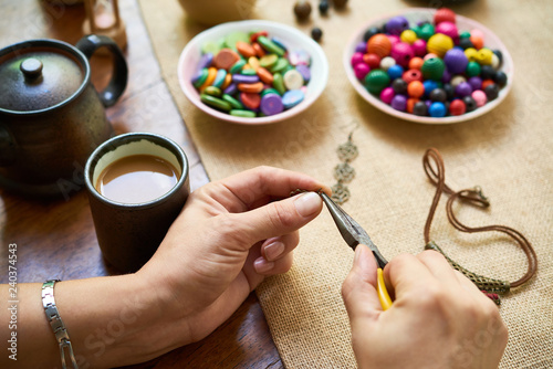 Close-up of woman sitting at the table drinking coffee and making handmade accessories from colorful beads