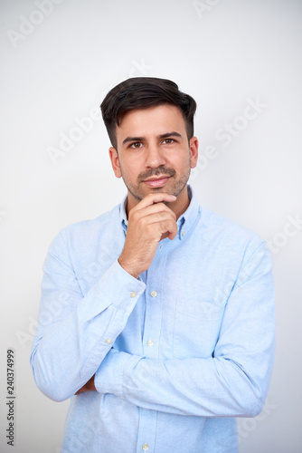 Portrait of young pensive man with dark hair and blue shirt standing on white background