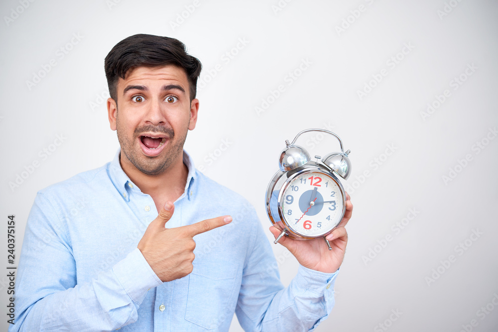 Shocked man standing on white background and pointing at alarm clock because its deadline Shocked man standing on white background and pointing at alarm clock because its deadline