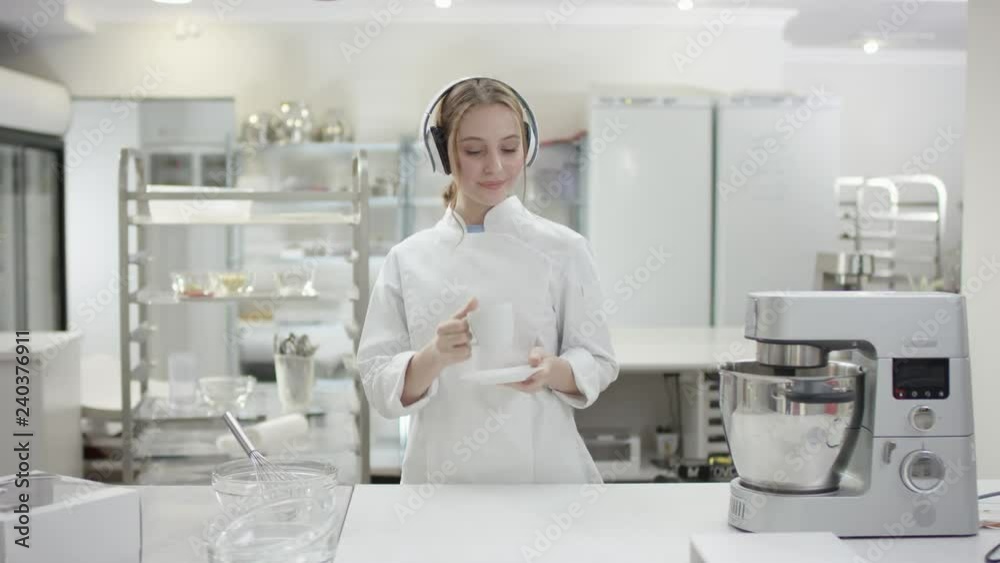 Young lady sous chef is having breakfast at professional kitchen in uniform and earphones Young lady sous chef is having breakfast at professional kitchen in uniform and earphones