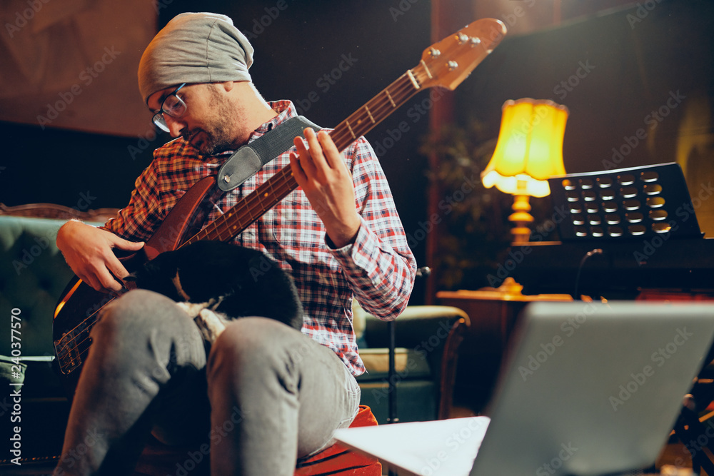 Obraz premium Young Caucasian man with hat on head playing bass guitar while sitting in home studio.