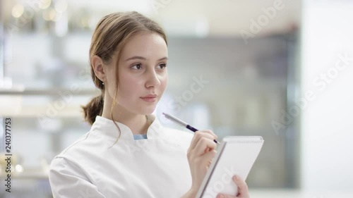 Young lady chef looking at camera, smiling chef in chef's form on by professional restaurant kitchen. Young woman is writing a menu