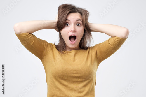 Shocked amazed young woman with hands on head standing and shouting. Studio shoot