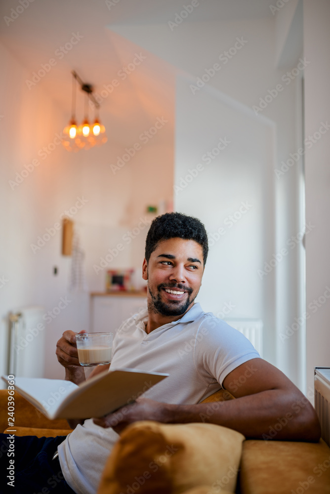 Fototapeta premium Handsome young man sitting on sofa, reading a book and drinking coffee.