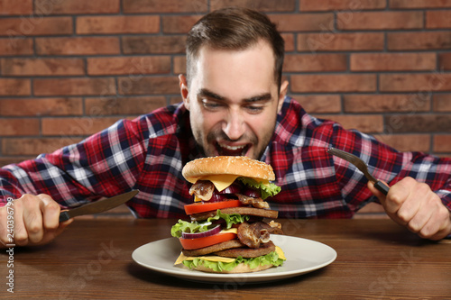 Young hungry man with cutlery eating huge burger at table