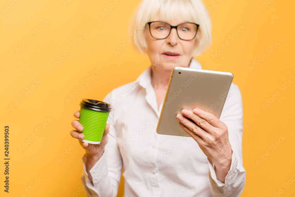Happy senior woman using tablet while drinking coffee isolated over yellow background. Happy senior woman using tablet while drinking coffee isolated over yellow background.