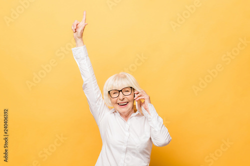 Portrait of a cheerful senior woman gesturing victory isolated over yellow background. Using phone.