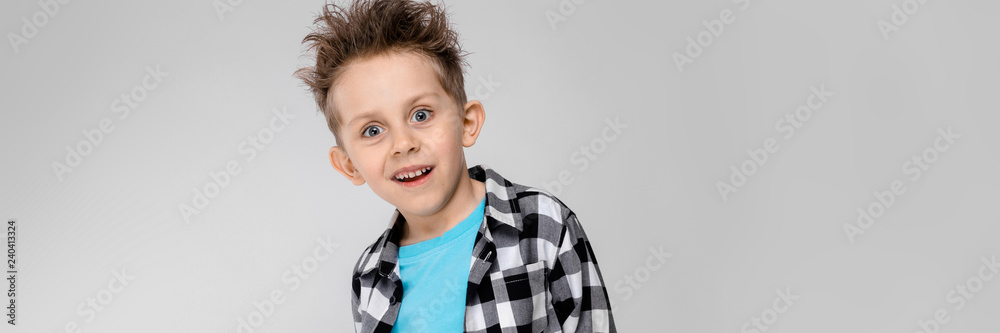 A handsome boy in a plaid shirt, blue shirt and jeans stands on a gray background. The boy folded his arms behind his back A handsome boy in a plaid shirt, blue shirt and jeans stands on a gray background. The boy folded his arms behind his back