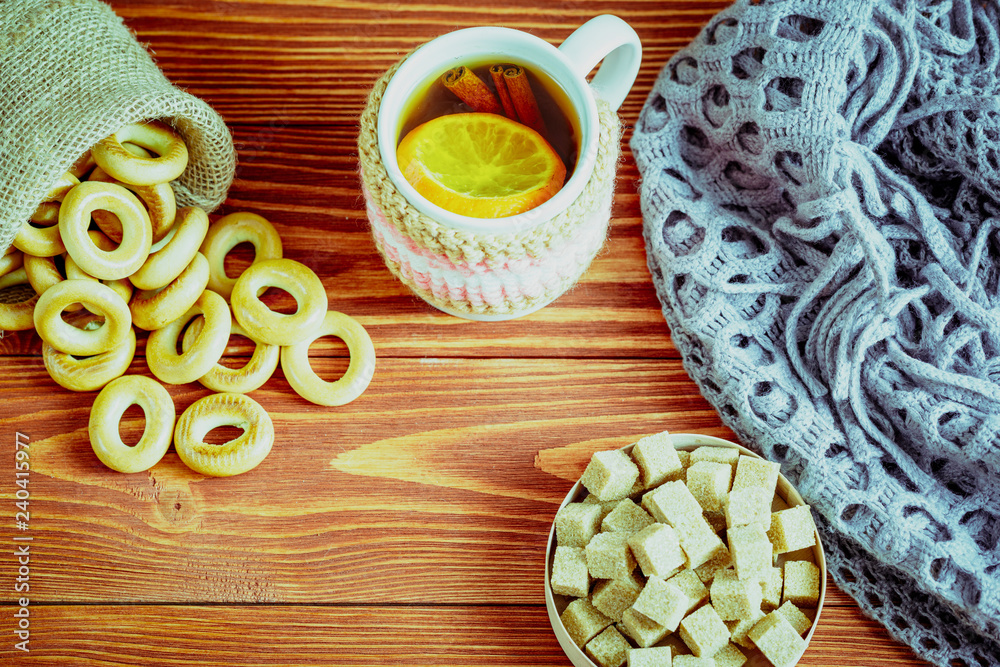 A cup of tea and a woolen scarf. There is a orange and two cinnamon sticks in the cup. With bagels in a bag and brown sugar. Top view. Space for text. A cup of tea and a woolen scarf. There is a orange and two cinnamon sticks in the cup. With bagels in a bag and brown sugar. Top view. Space for text.