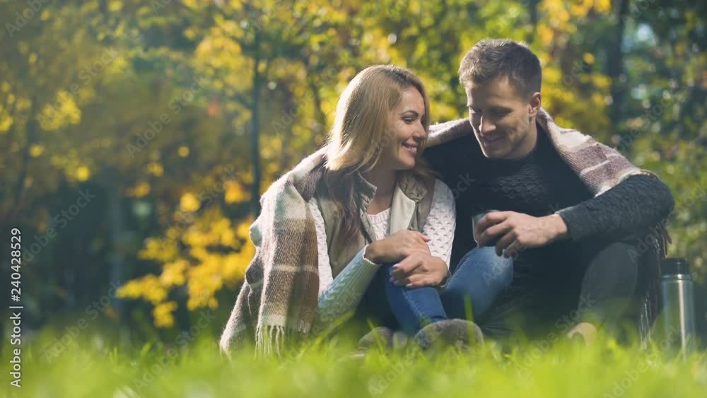 Couple covered with plaid sitting in autumn park, drinking hot tea and speaking Couple covered with plaid sitting in autumn park, drinking hot tea and speaking