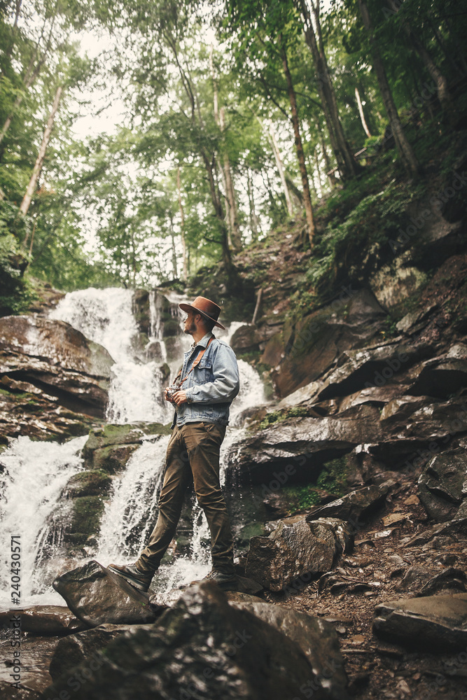 Stylish young man in hat traveling in mountains. Hipster man with photo camera exploring  beautiful waterfall in summer forest. Travel and wanderlust concept. Space for text.