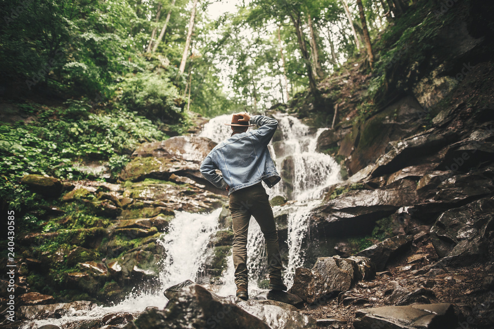 Travel and wanderlust concept. Stylish young man in hat traveling in mountains. Hipster man looking at beautiful waterfall in summer forest.  Space for text.