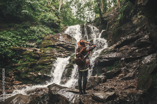 Stylish hipster girl in hat traveling in mountains. Young woman with backpack exploring in summer forest  looking at waterfall. Travel and wanderlust concept. Space for text