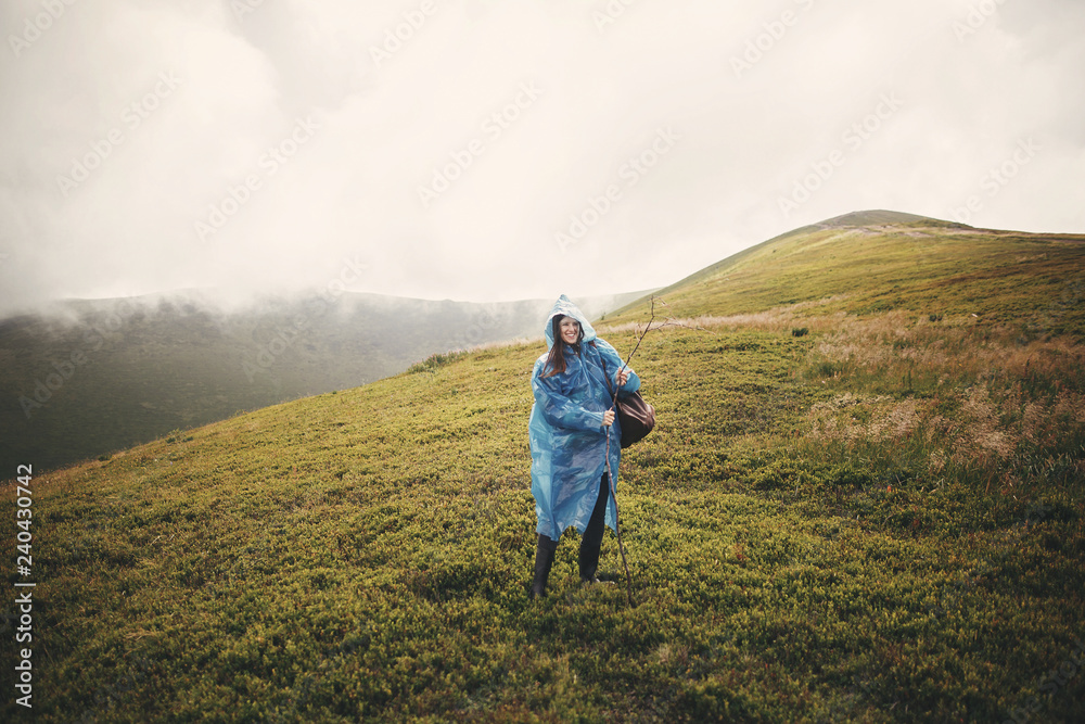 Stylish hipster girl in blue raincoat and with backpack standing on top of misty mountains and clouds. Funny young woman traveler smiling and exploring mountains. Travel and wanderlust