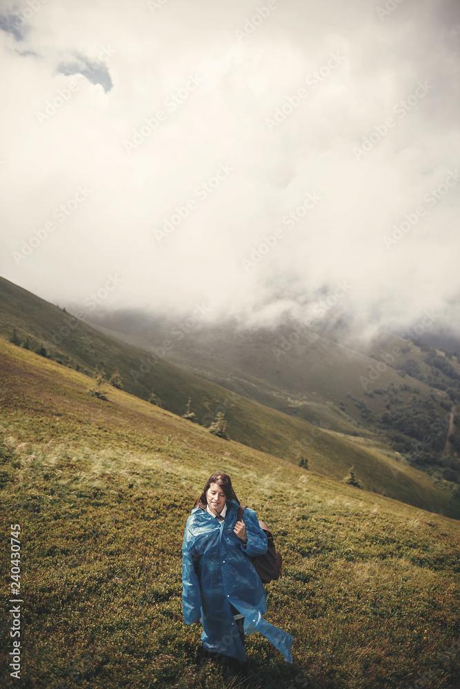Stylish hipster girl in blue raincoat and with backpack standing on top of misty mountains and clouds. Young woman traveler exploring mountains. Travel and wanderlust concept. Atmospheric moment