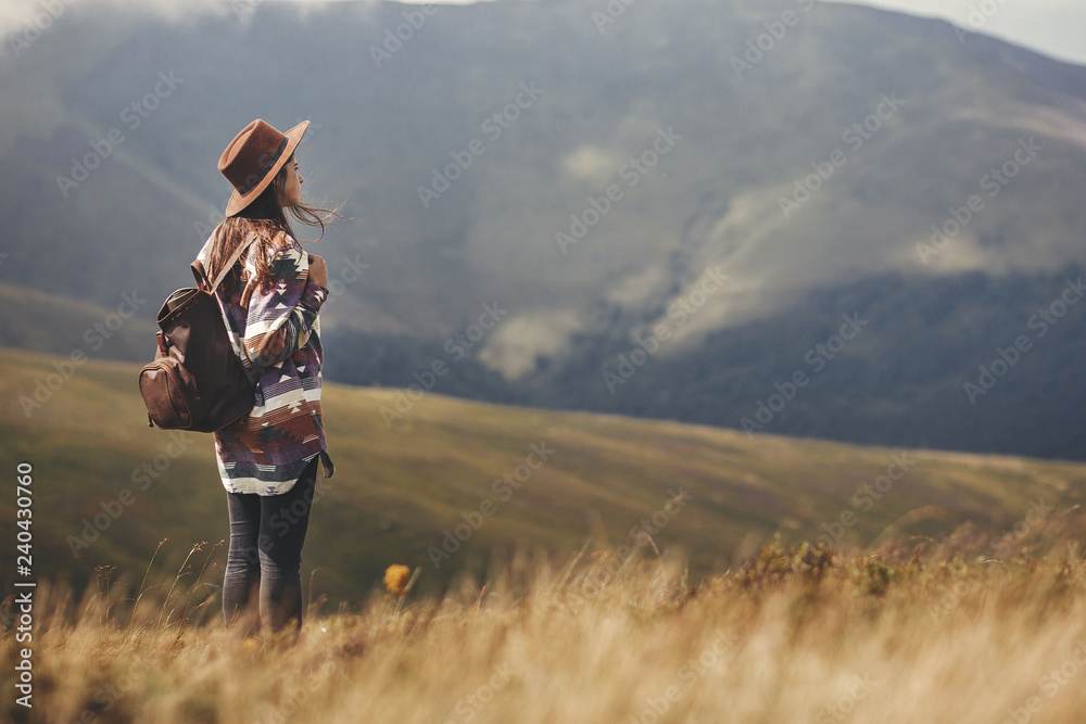 Stylish hipster girl in hat walking on top of mountains. Happy young woman with backpack exploring sunny mountains. Travel and wanderlust concept. Amazing atmospheric moment - obrazy, fototapety, plakaty 