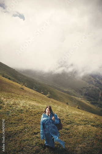 Stylish hipster girl in blue raincoat and with backpack standing on top of misty mountains and clouds. Young woman traveler exploring mountains. Travel and wanderlust concept. Atmospheric moment