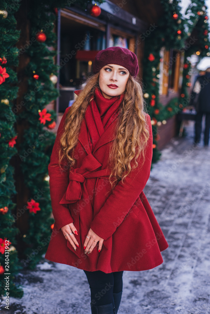 A beautiful girl in a red coat and a beret is walking along the Christmas street.
