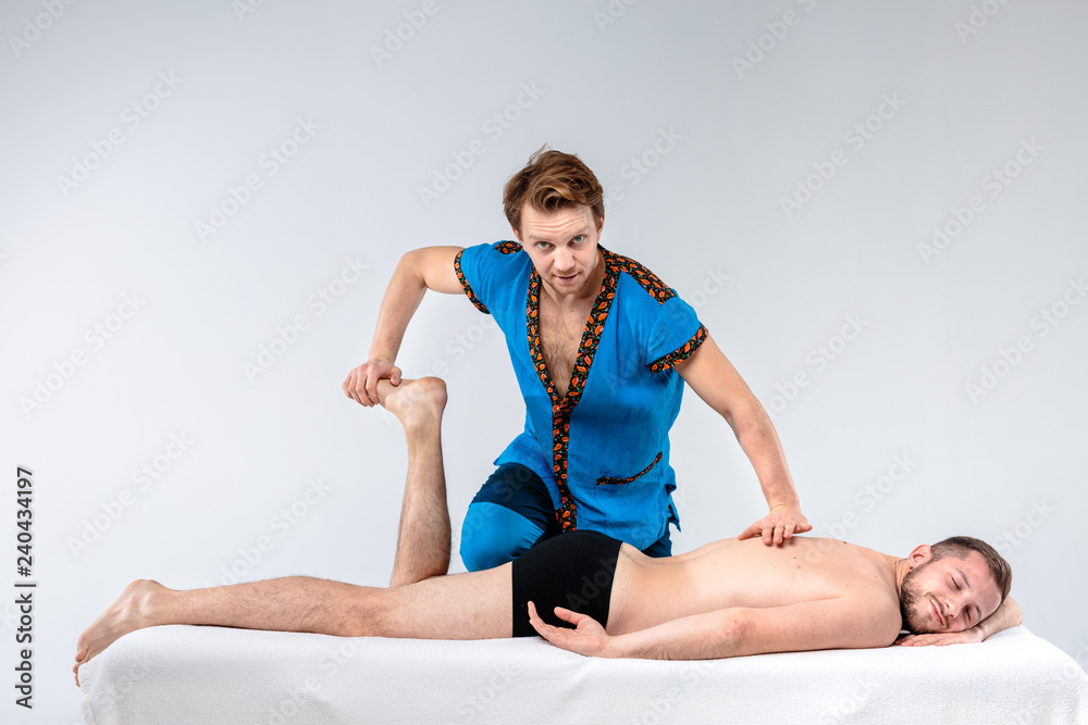 Theme massage and body care. A handsome Caucasian man in blue uniform and beard does stretching, heals, the diagnosis of muscles of legs and back to bare-footed guy with good figure on massage table Theme massage and body care. A handsome Caucasian man in blue uniform and beard does stretching, heals, the diagnosis of muscles of legs and back to bare-footed guy with good figure on massage table