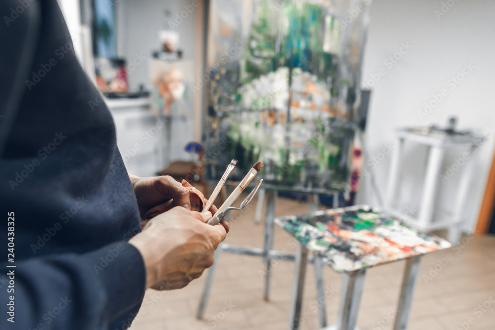 Painter holds a brush and a palette knife in the background of the easel with the host and a palette of oil paints. Artist's hands with brushes close-up on a background of artistic studio.Oil painting