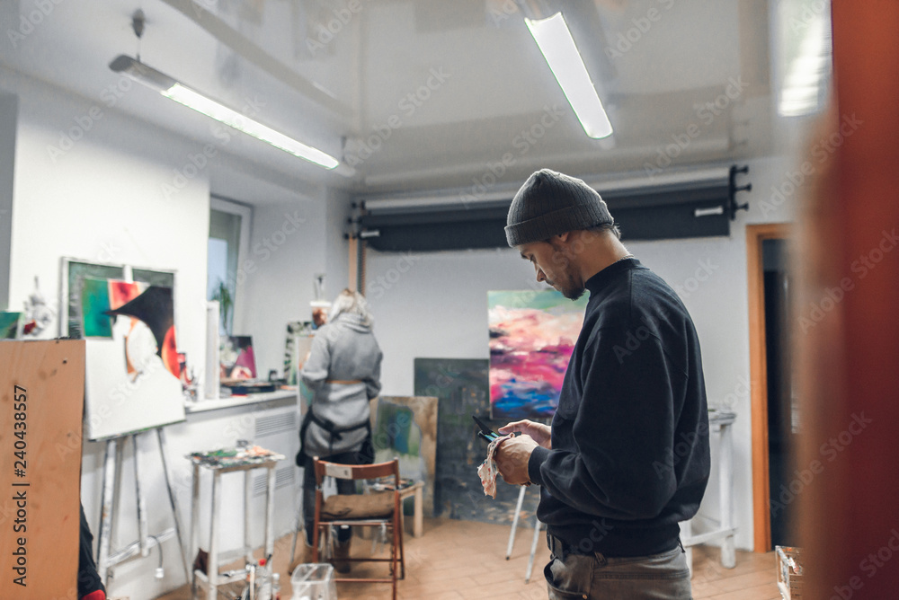 Portrait of a young artist standing in the studio and using the internet on a smartphone,against the background of paintings,easels and the painter paints a picture.Creative atmosphere in art studio