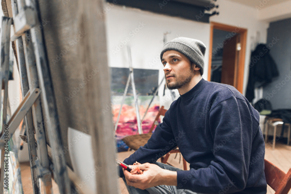 Portrait of a contemporary young man painting a picture at a workshop in painting, holding a brush in his hands and looking at the host. Painter creates a painting by the house in the studio.