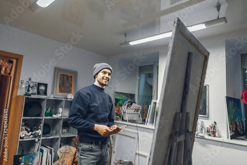 Smiling young artist stands with a brush and palette knife in the hands of the studio, looking at the picture that he paints and smiles. Positive artist paints a picture in a cozy studio.