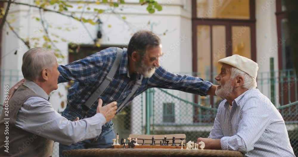 Two Caucasian old men playing chess game in the yard and their friend coming to them, greeting and shaking hands. Outdoor. Two Caucasian old men playing chess game in the yard and their friend coming to them, greeting and shaking hands. Outdoor.