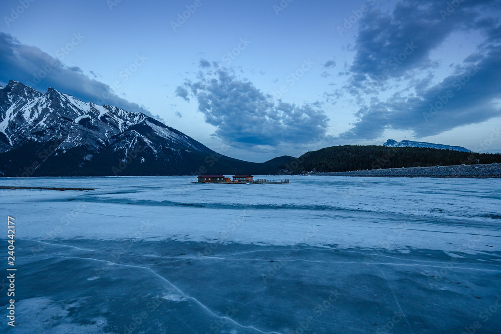 Lake Minnewanka Banff, Canada. Glacial lake completely frozen and covered in snow. Blue hour at the lake with glacial mountain background. Cabin on the frozen lake.