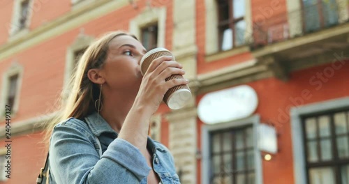 View from the side on the young pretty Caucasian girl in the jeans jacket walking the street with nice architecture, scrolling ot tapping pn the smartphonr and drinking coffee. Close up. Outdoor. photo