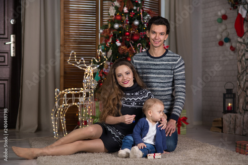 family sits with the baby sitting near the Christmas tree, © Evgenia Tiplyashina