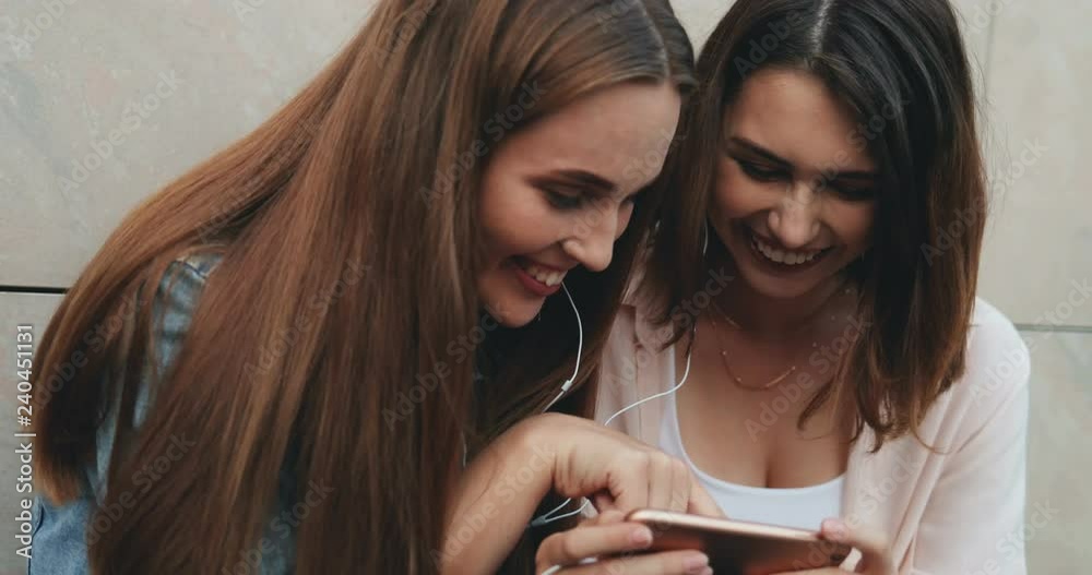 Close up of the two cheerful attractive young Caucasian women best friends in headphones laughing a lot when watching something on the smartphone screen. Outside. Close up of the two cheerful attractive young Caucasian women best friends in headphones laughing a lot when watching something on the smartphone screen. Outside.