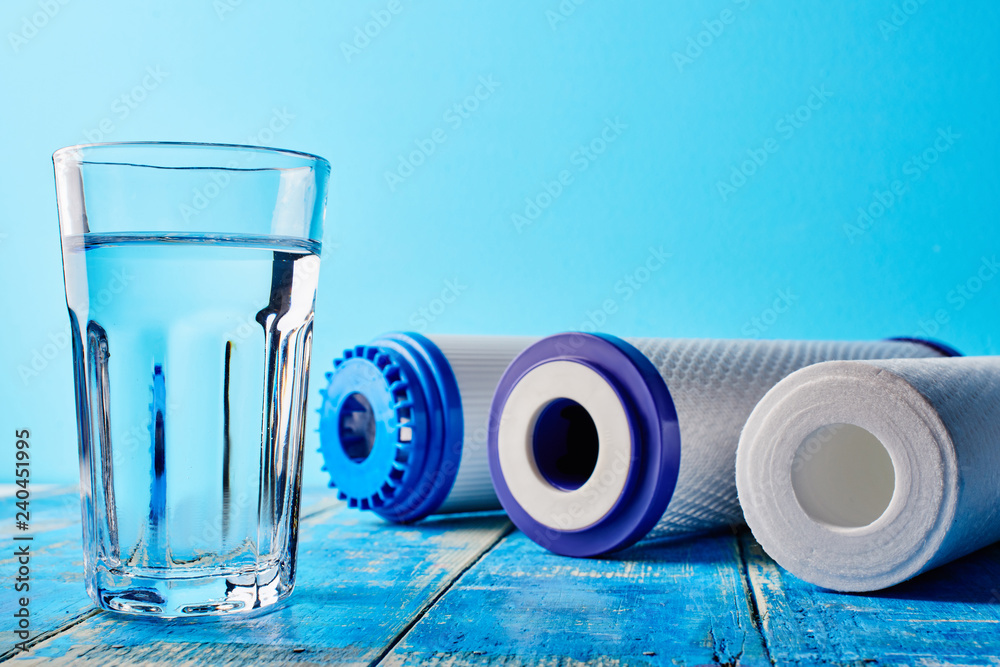 Water filters. Carbon cartridges and a glass on a blue background. Household filtration system. - obrazy, fototapety, plakaty 