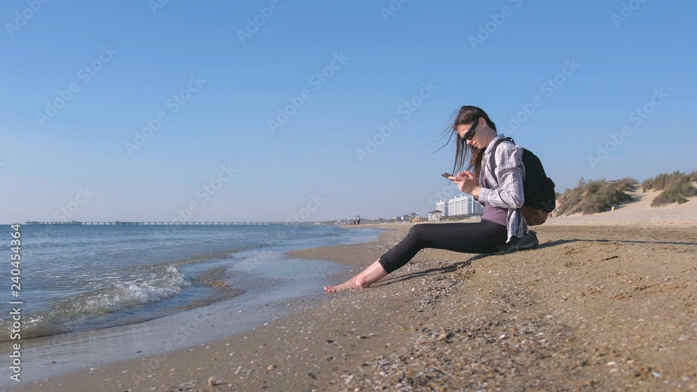 Traveler girl with a backpack sitting on a sandy sea beach and typing a message on mobile phone.