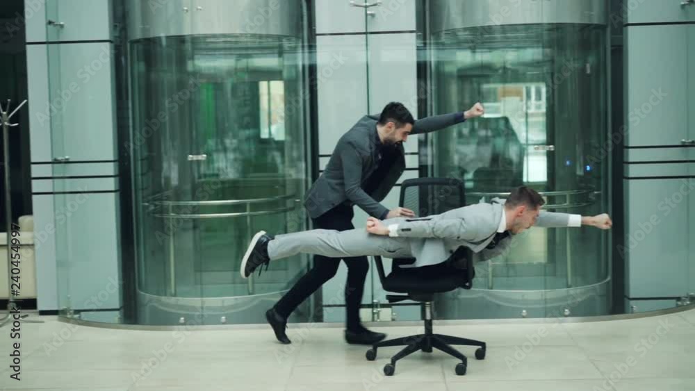 Funny young men are having fun with rolling chair, one is pushing armchair other is lying in it and moving forward in office hall. Youth and joy concept.