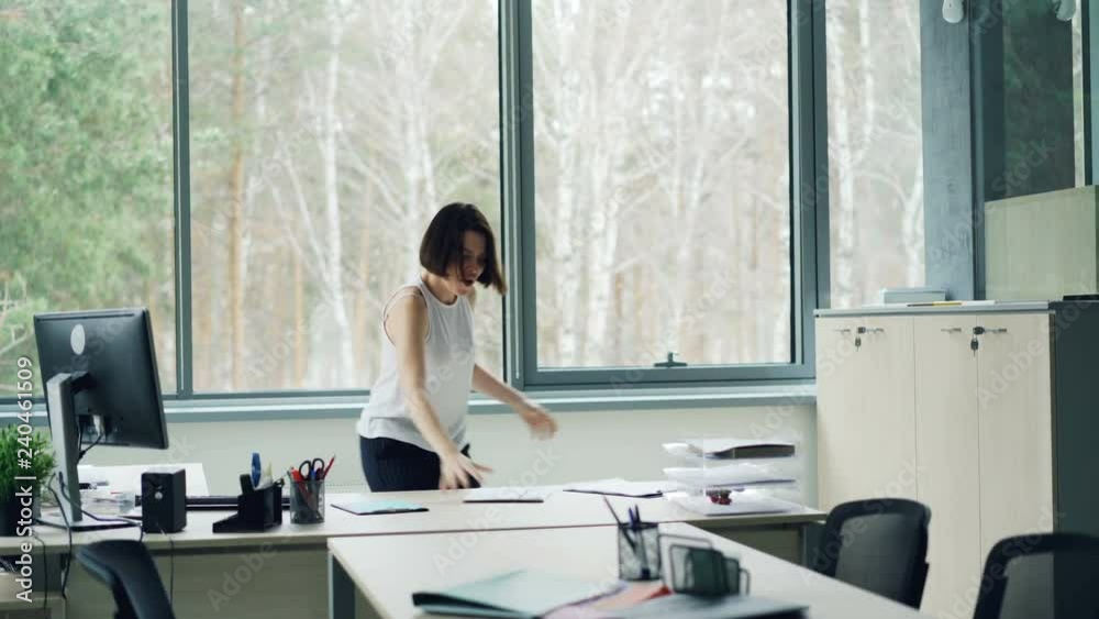 Happy young woman is dancing in office taking off jacket and throwing papers tossing her hair enjoying freedom and good mood. Corporate fun and youth concept.