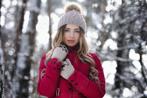 Beautiful girl with long hair in white knitted hat having fun outdoor in winter forest under snowflakes. Pretty young model standing on forest background and looking at camera. - Image