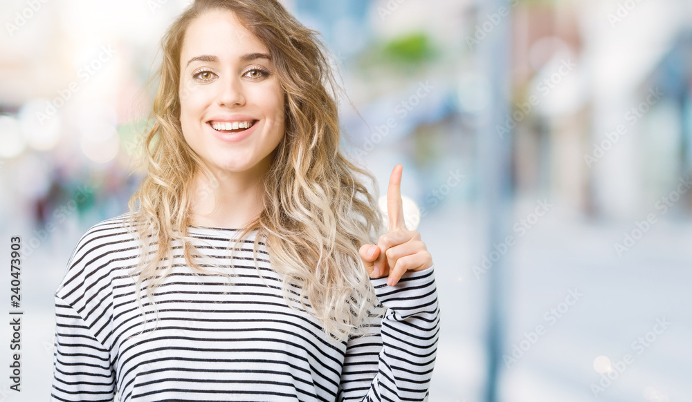 Beautiful young blonde woman wearing stripes sweater over isolated background showing and pointing up with finger number one while smiling confident and happy.