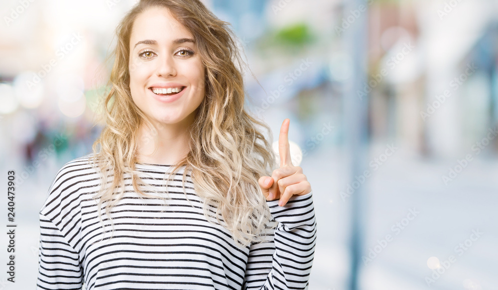 Beautiful young blonde woman wearing stripes sweater over isolated background showing and pointing up with finger number one while smiling confident and happy. Beautiful young blonde woman wearing stripes sweater over isolated background showing and pointing up with finger number one while smiling confident and happy.