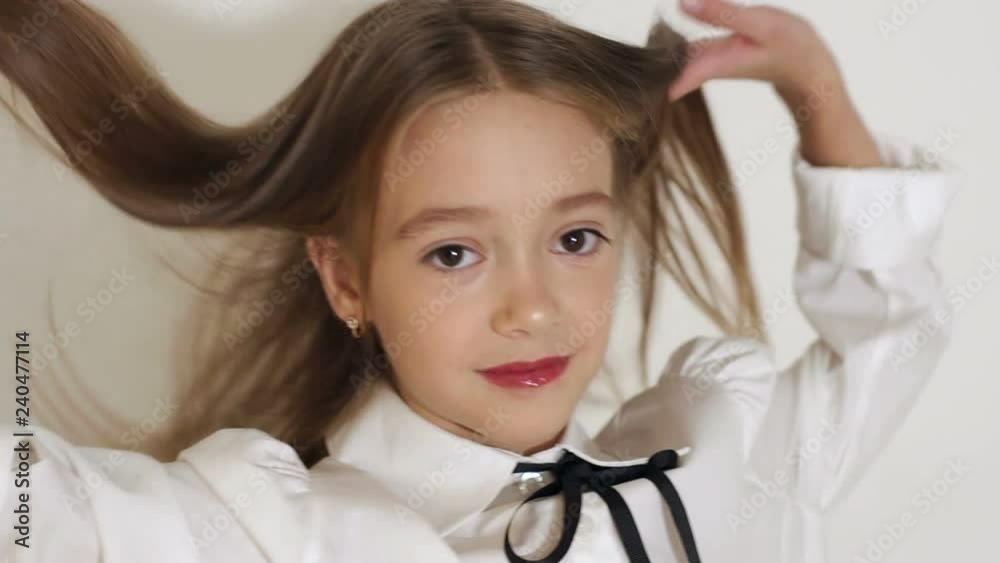 Close-up portrait of a cute little girl with big brown eyes and long hair posing in front of the camera in a studio with a white wall, she looks into the camera and throws up her hair with her hands.