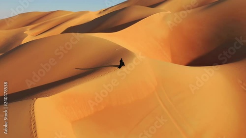  Aerial view from a drone flying next to a woman in abaya (United Arab Emirates traditional dress) walking on the dunes in the desert. Dubai, UAE. photo