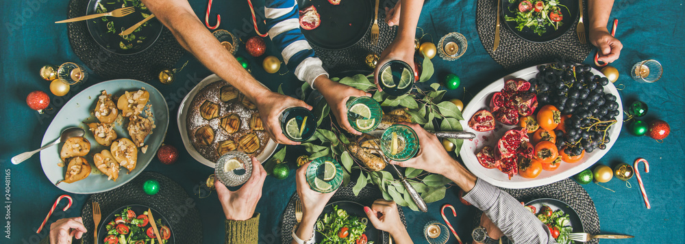 Company of friends gathering for Christmas or New Year party dinner at festive table. Flat-lay of human hands holding glasses with drinks, celebrating holiday together, top view, wide composition