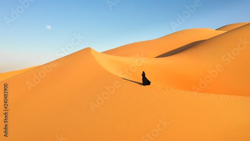 Aerial view from a drone flying next to a woman in abaya (United Arab Emirates traditional dress) walking on the dunes in the desert of the Empty Quarter. Abu Dhabi, UAE.