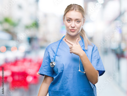 Fototapeta Naklejka Na Ścianę i Meble -  Young blonde surgeon doctor woman over isolated background cheerful with a smile of face pointing with hand and finger up to the side with happy and natural expression on face looking at the camera.