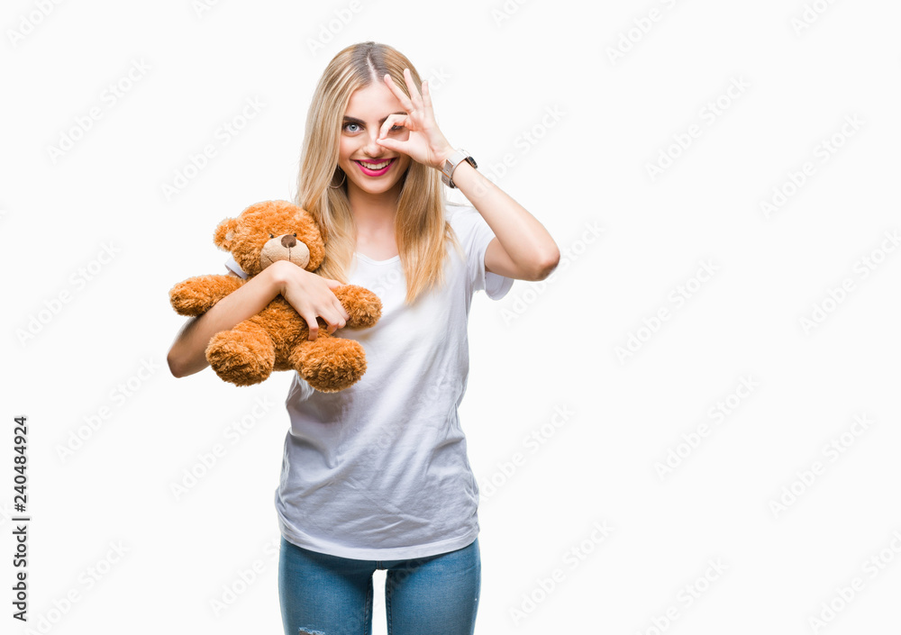 Young beautiful blonde woman holding teddy bear over isolated background with happy face smiling doing ok sign with hand on eye looking through fingers Young beautiful blonde woman holding teddy bear over isolated background with happy face smiling doing ok sign with hand on eye looking through fingers