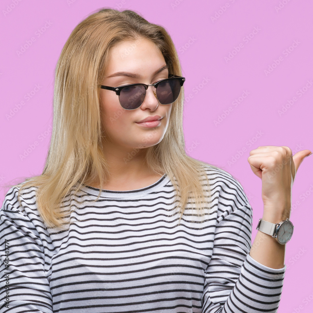 Young caucasian woman wearing sunglasses over isolated background smiling with happy face looking and pointing to the side with thumb up. Young caucasian woman wearing sunglasses over isolated background smiling with happy face looking and pointing to the side with thumb up.